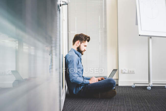 Jovem do sexo masculino sentado no chão do escritório usando laptop — Fotografia de Stock