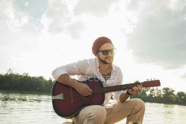 Jovem sentado junto ao lago tocando guitarra — Fotografia de Stock