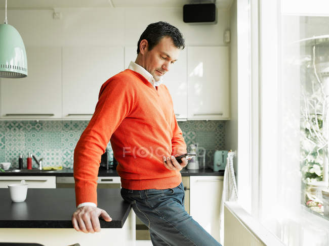 Зрелый мужчина использует мобильный телефон на кухне — стоковое фото