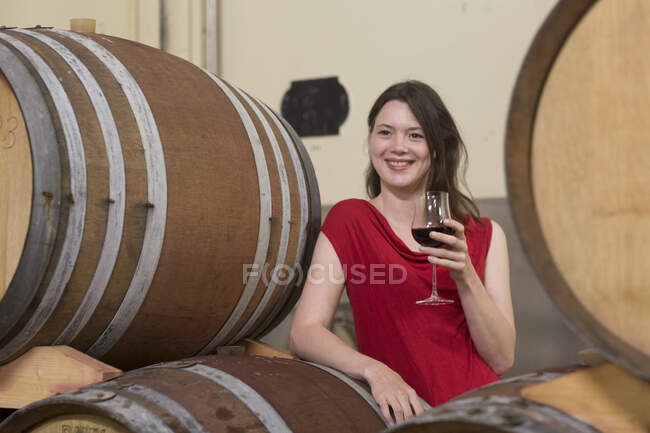 Portrait of young woman in wine cellar, leaning against wine barrel, holding glass of wine — Stock Photo