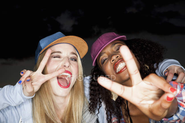 Young woman wearing baseball cap arm around friend looking at camera doing peace sign — Stock Photo