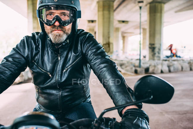 Mature male motorcyclist wearing black helmet and goggles sitting on motorcycle under flyover — Stock Photo