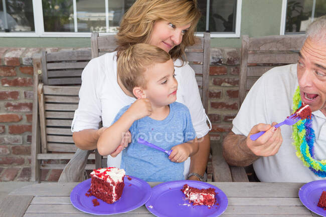 Nieto y madre viendo hombre mayor comer pastel de cumpleaños - foto de stock