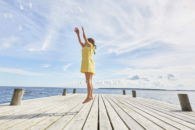 Молодая девушка на деревянном пирсе, прыгая, чтобы достичь пузырьков — стоковое фото