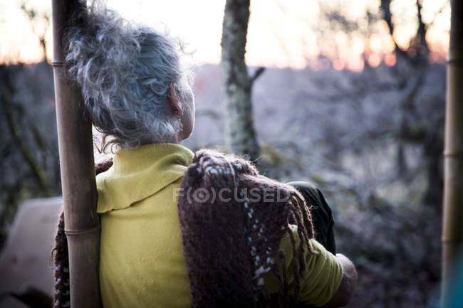 Вид ззаду зрілої жінки, що сидить на лісовому ганку, спостерігаючи за заходом сонця — стокове фото