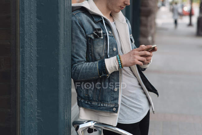 Молодий чоловік спирається на стіну, використовуючи смартфон, середній розділ — стокове фото