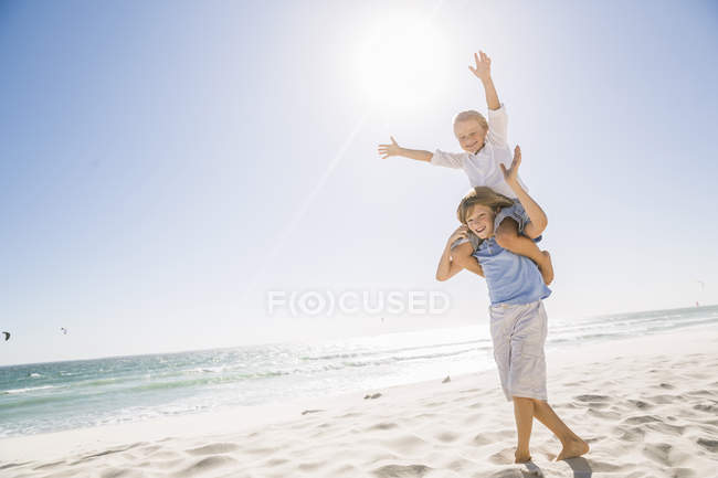 Vista completa del hermano mayor en la playa llevando al hermano sobre los hombros, los brazos levantados sonriendo - foto de stock