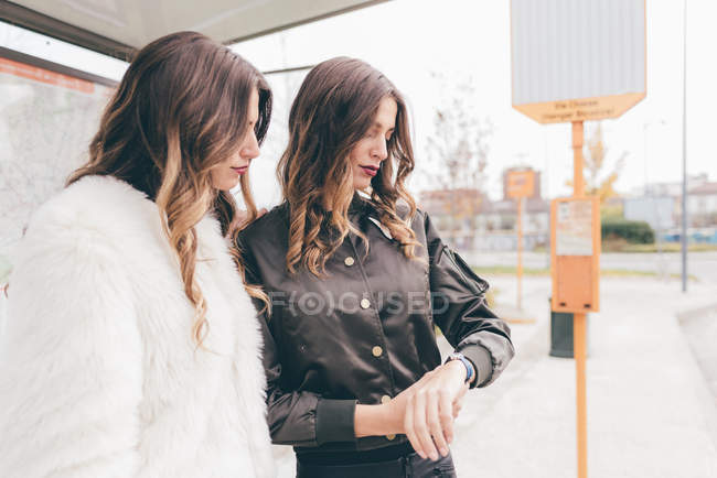 Twin sisters standing at bus shelter, looking at wristwatch — Stock Photo