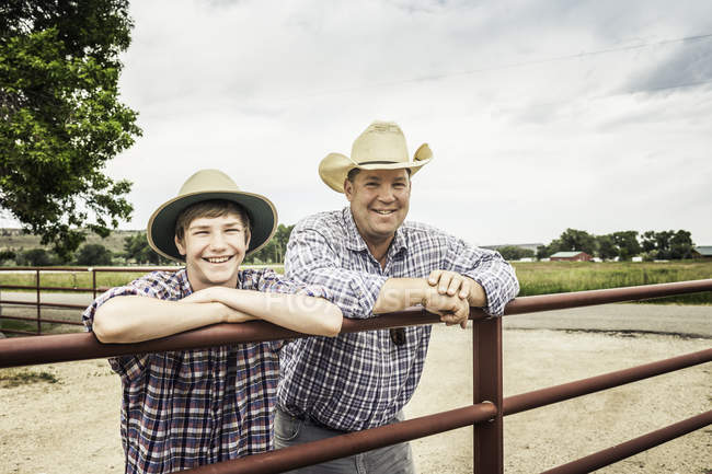 Portrait of farmer and teenage grandson leaning against gate — Stock Photo