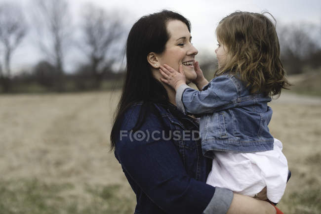 Madre sosteniendo a su hija pequeña, al aire libre, cara a cara, sonriendo - foto de stock