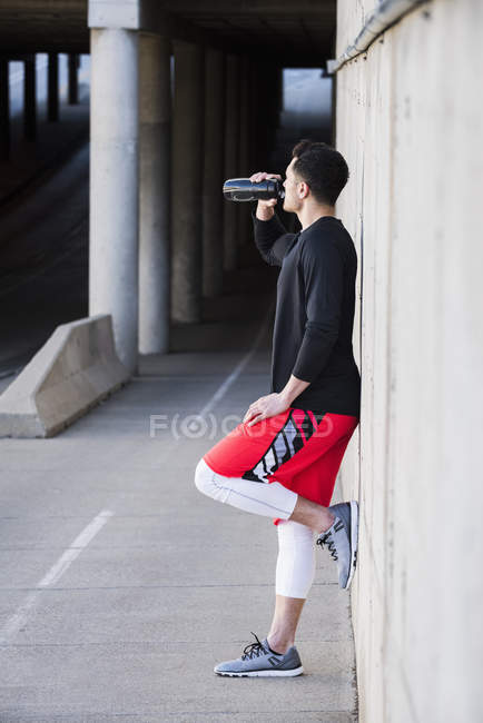 Young male runner leaning against underpass wall drinking water — Stock Photo