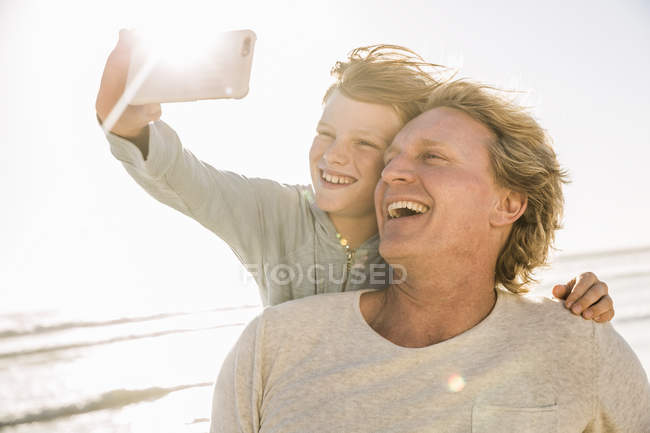 Filho na praia usando smartphone para tirar selfie sorrindo — Fotografia de Stock
