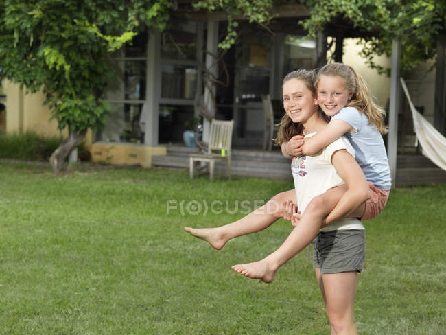 Adolescente menina dando irmã piggyback passeio no jardim — Fotografia de Stock