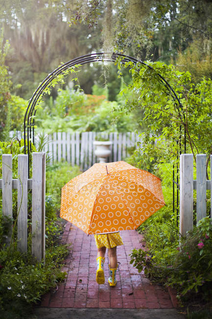 Vista trasera de la niña caminando a través de la puerta del jardín que lleva paraguas - foto de stock