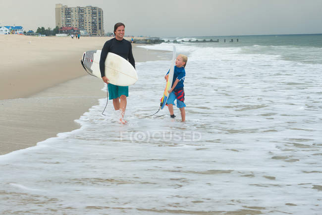 Père et fils marchant le long de la plage, portant des planches de surf — Photo de stock