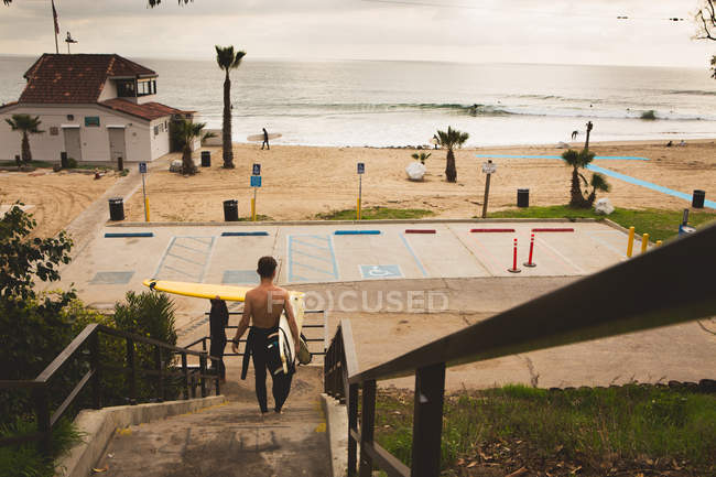 Giovane uomo che cammina verso la spiaggia, portando la tavola da surf, vista posteriore — Foto stock