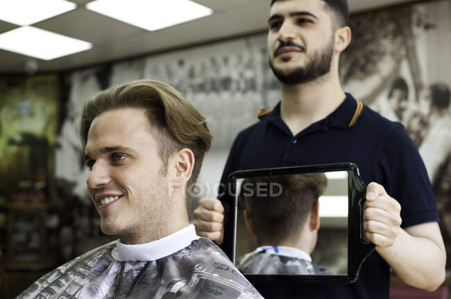 Barbiere utilizzando specchio per mostrare taglio di capelli cliente sorridente — Foto stock