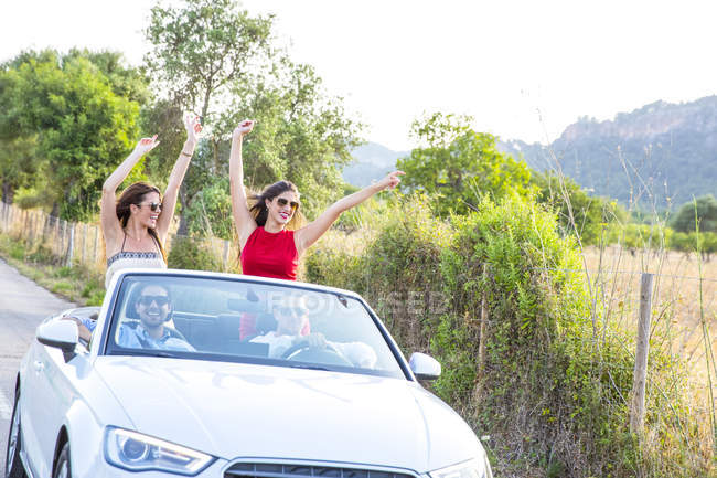 Two young female friends standing waving whilst driving on rural road in convertible, Majorca, Spain — Stock Photo
