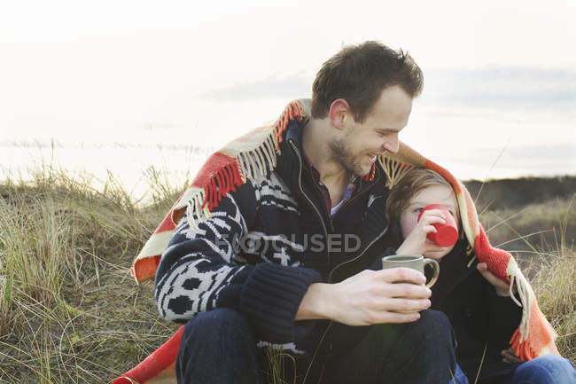 Smiling mid adult man and son wrapped in blanket on sand dunes — Stock Photo