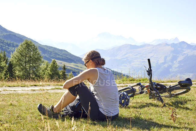 Feminino mountain biker sentado na paisagem de montanha ler textos de smartphones, Vale de Aosta, Aosta, Itália — Fotografia de Stock