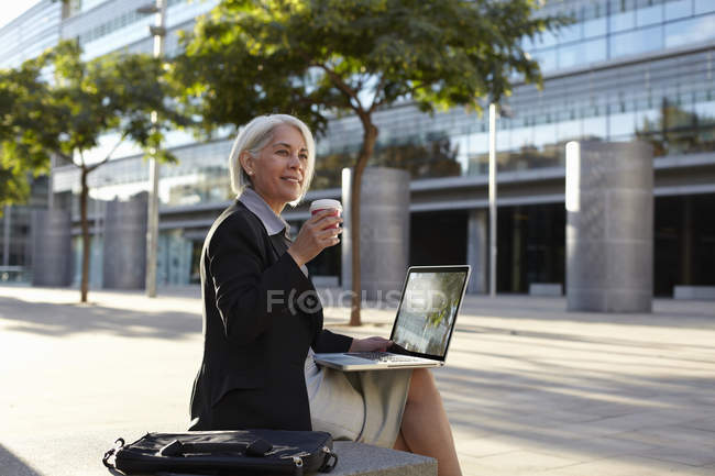 Empresária sentada fora usando laptop e bebendo bebida quente — Fotografia de Stock