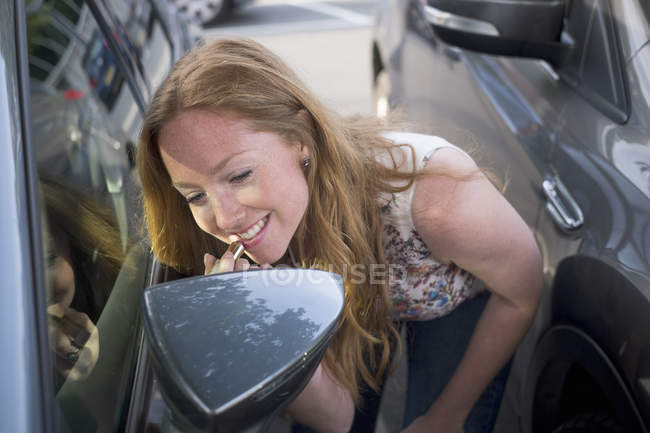 Jeune femme appliquant rouge à lèvres tout en regardant dans le miroir de vent de voiture — Photo de stock