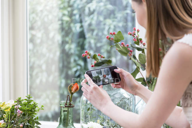Sopra la spalla vista della donna utilizzando lo smartphone per scattare foto di fiori in vaso — Foto stock
