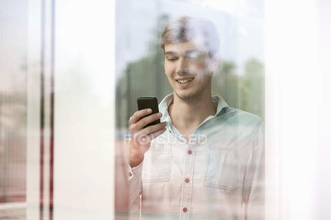 Young man behind reflective glass looking at smartphone — Stock Photo