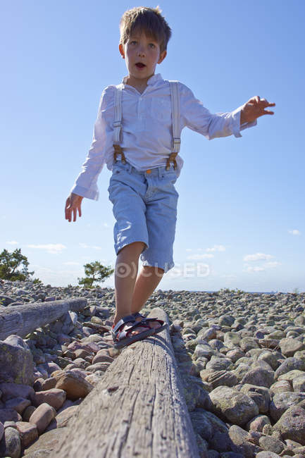Мальчик балансирует на бревне на пляже с камнями — стоковое фото