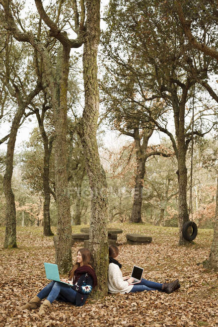 Sisters using laptop on fallen leaves under trees in autumn forest — Stock Photo