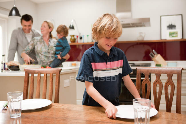 Filho ajudando a colocar a mesa com os pais e irmão visível atrás na cozinha — Fotografia de Stock