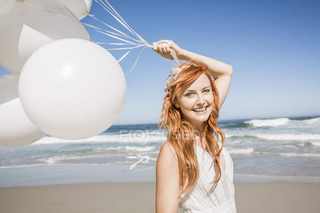 Mujer pelirroja en la playa sosteniendo globos mirando a la cámara sonriendo - foto de stock