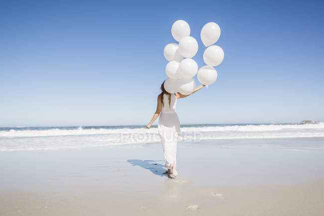 Vista trasera de longitud completa de la mujer en la playa con vestido blanco celebración de globos - foto de stock