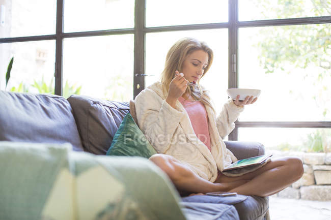 Woman sitting on sofa eating summer fruits and using digital tablet — Stock Photo