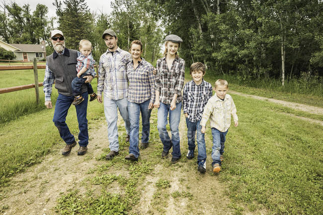 Vista frontal de longitud completa de la familia de varias generaciones caminando lado a lado en la pista de tierra mirando a la cámara sonriendo - foto de stock