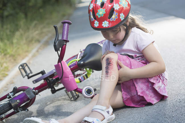 Little girl with injured leg sitting on road with bicycle — Stock Photo