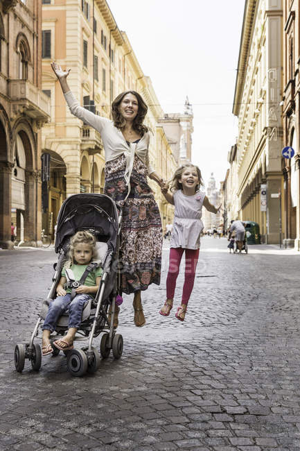 Mother and daughter jumping in street while pushing baby carriage, Bologna, Italy — Stock Photo