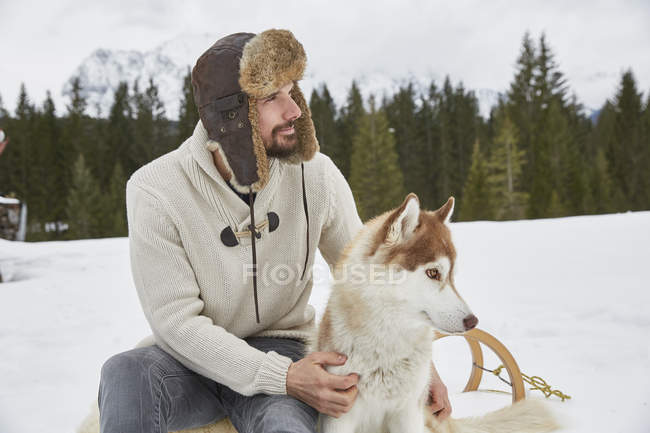 Jovem usando chapéu de caçador acariciando husky na neve, Elmau, Baviera, Alemanha — Fotografia de Stock