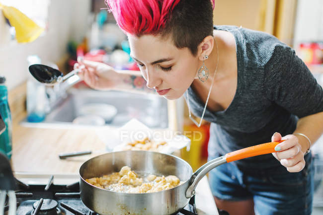 Mujer joven con cabello rosa oliendo comida frita en la cocina - foto de stock