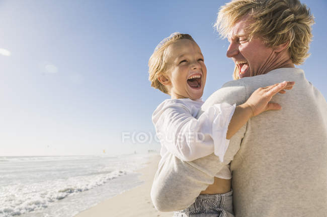 Father on beach carrying son, mouth open smiling — Stock Photo