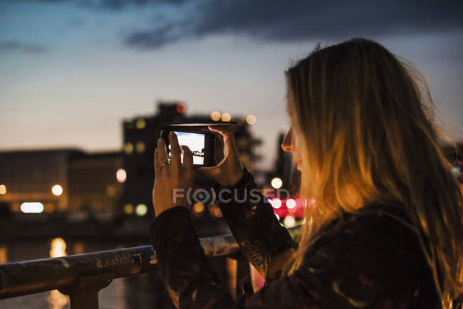 Young woman by waterfront, at dusk, photographing view with smartphone — Stock Photo