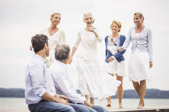 Groupe d'amis sur la jetée, amies marchant vers des amis masculins assis sur le bord de la jetée — Photo de stock