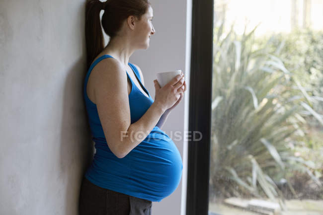 Side view of pregnant woman leaning against wall holding coffee cup looking away out of window smiling — Stock Photo