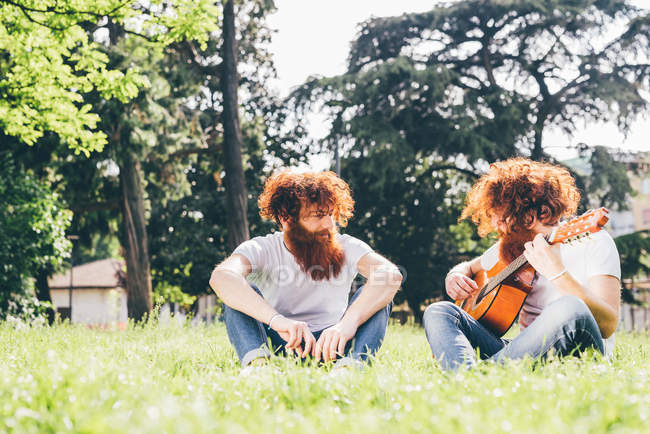 Молоді чоловіки хіпстер близнюки з червоними бородами сидять в парку граючи на гітарі — стокове фото