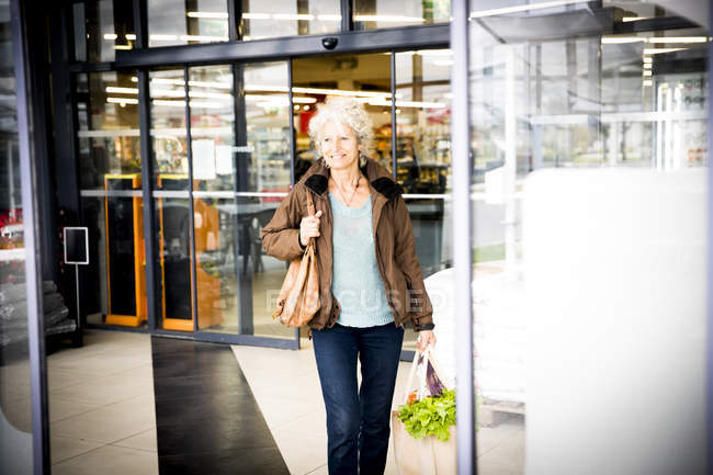 Mature woman exiting supermarket with bag of shopping — Stock Photo