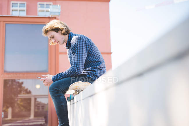 Молодой городской скейтбордист сидит на скейтборде и читает текст на смартфоне — стоковое фото