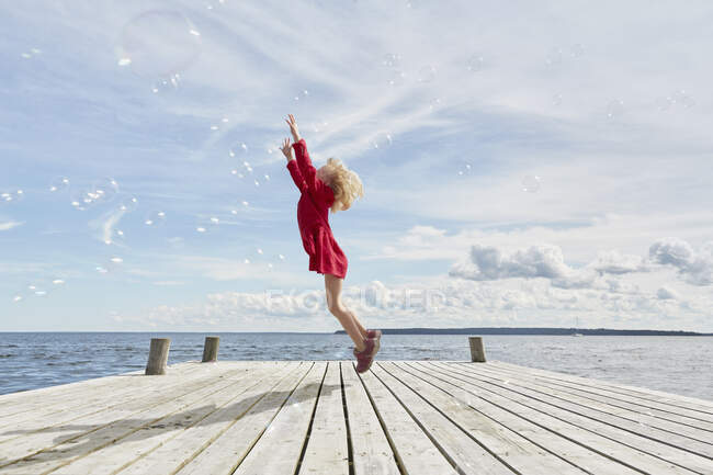 Young girl on wooden pier, jumping to reach bubbles — Stock Photo