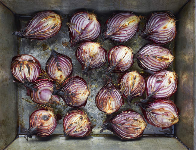 Overhead view of roasted red onion halves with rosemary and olive oil in roasting tin — Stock Photo