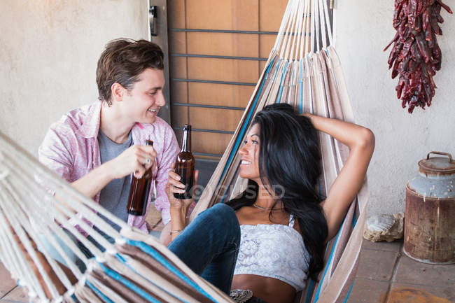 Pareja joven relajándose en la terraza, sosteniendo botellas de cerveza, mujer joven en hamaca - foto de stock