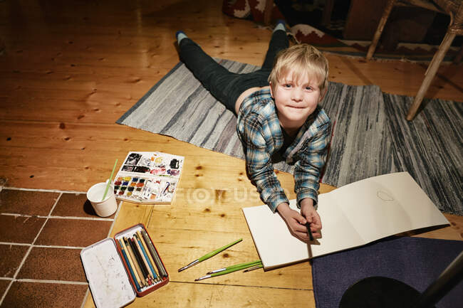 Хлопчик лежить на підлозі з акварельними фарбами та ескізним майданчиком — стокове фото
